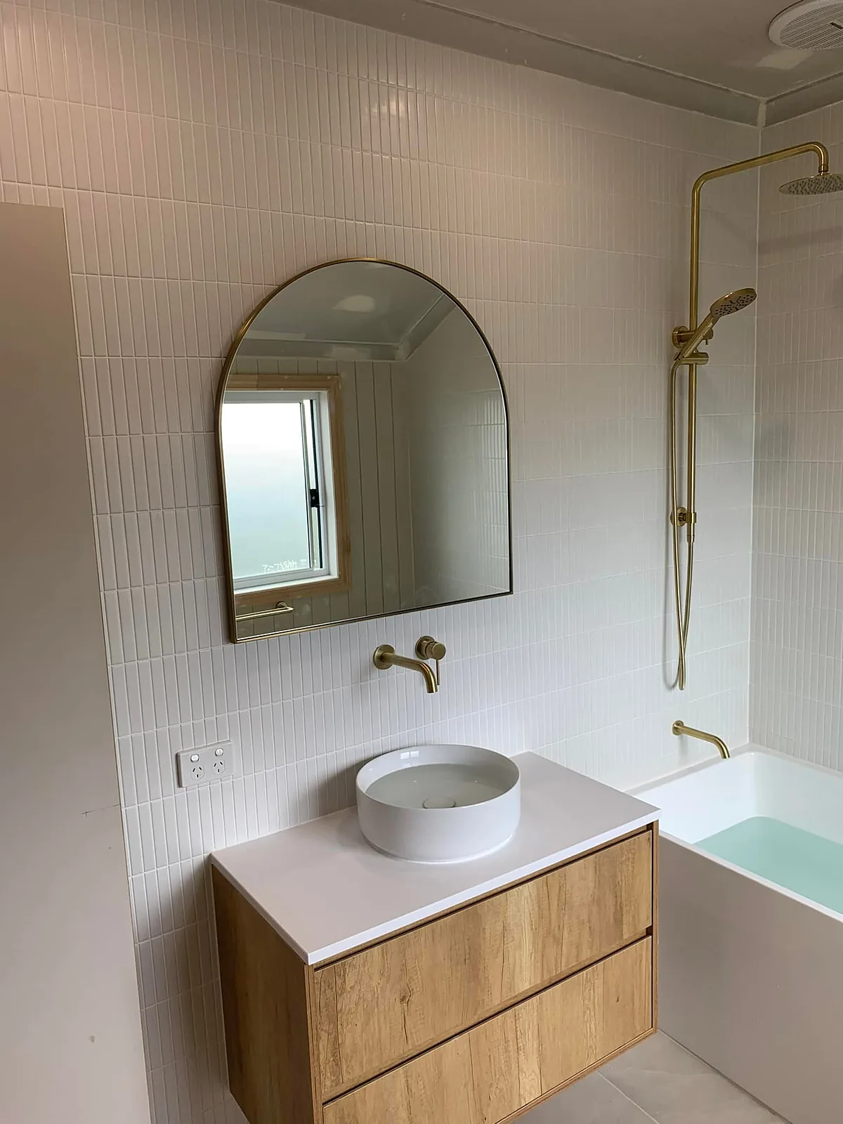 Finished bathroom with an arched mirror, round basin on a timber vanity and brushed brass shower and tapware