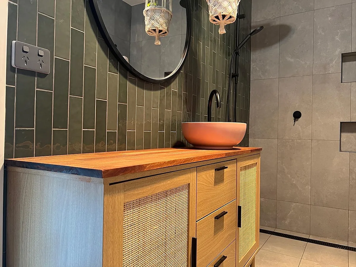 Green vertical tiles, a round black mirror and a timber and cane vanity with a terracotta basin